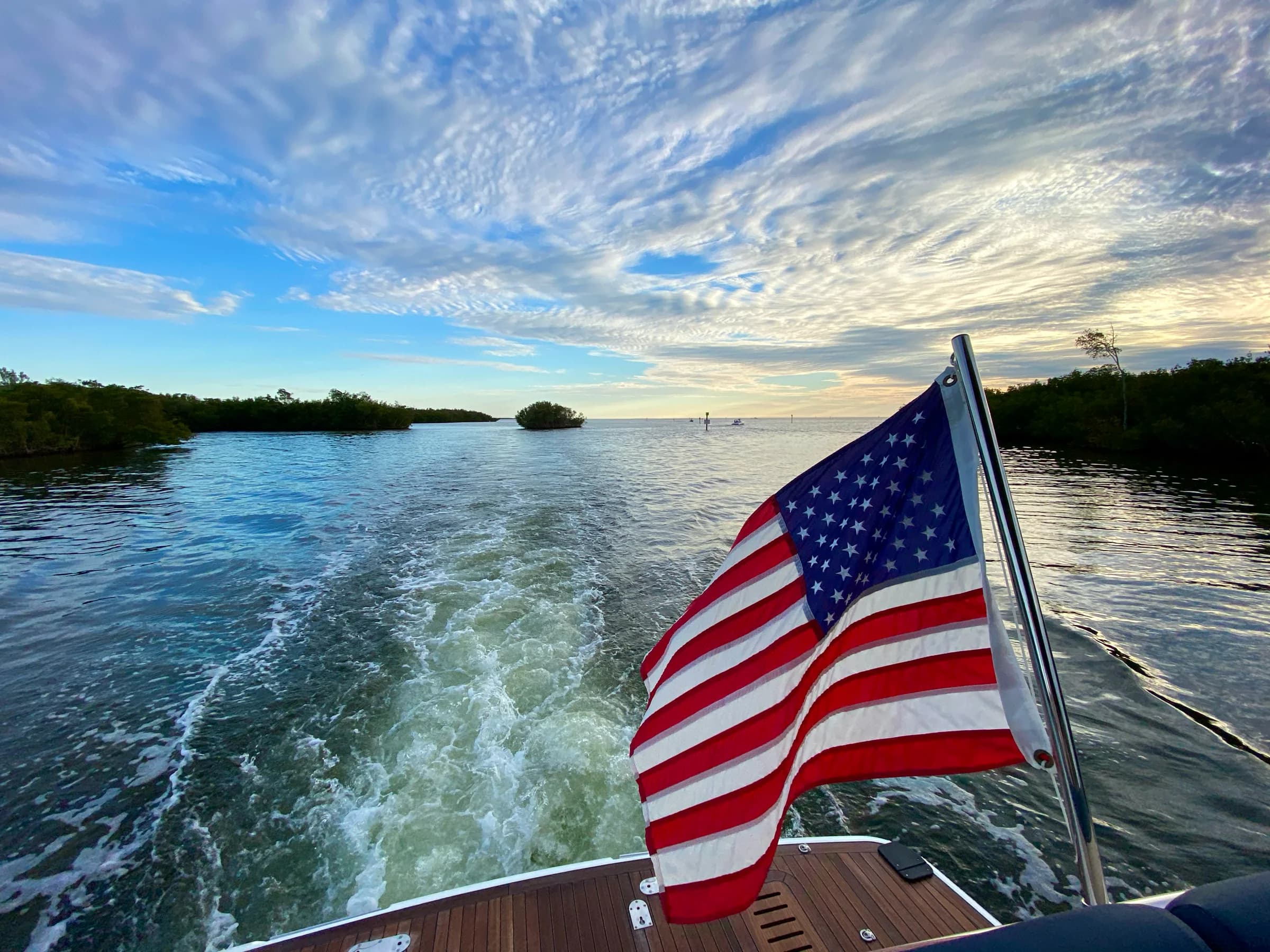 An American flag flying off a teak stern above the wake, mangrove islands and a sunset sky